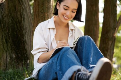 Portrait of happy asian girl sitting in park and writing in her diary. Young woman doing homework