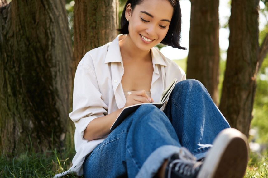 Portrait of happy asian girl sitting in park and writing in her diary. Young woman doing homework